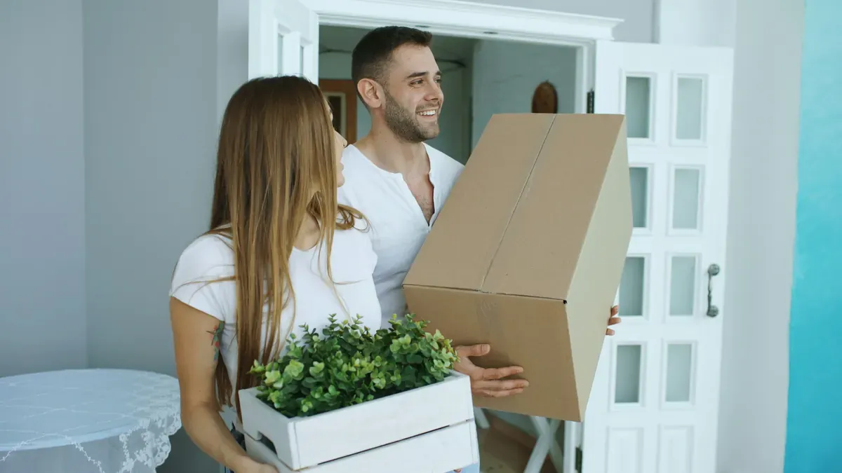Two people carrying a cardboard moving box and a potted plant through a doorway.