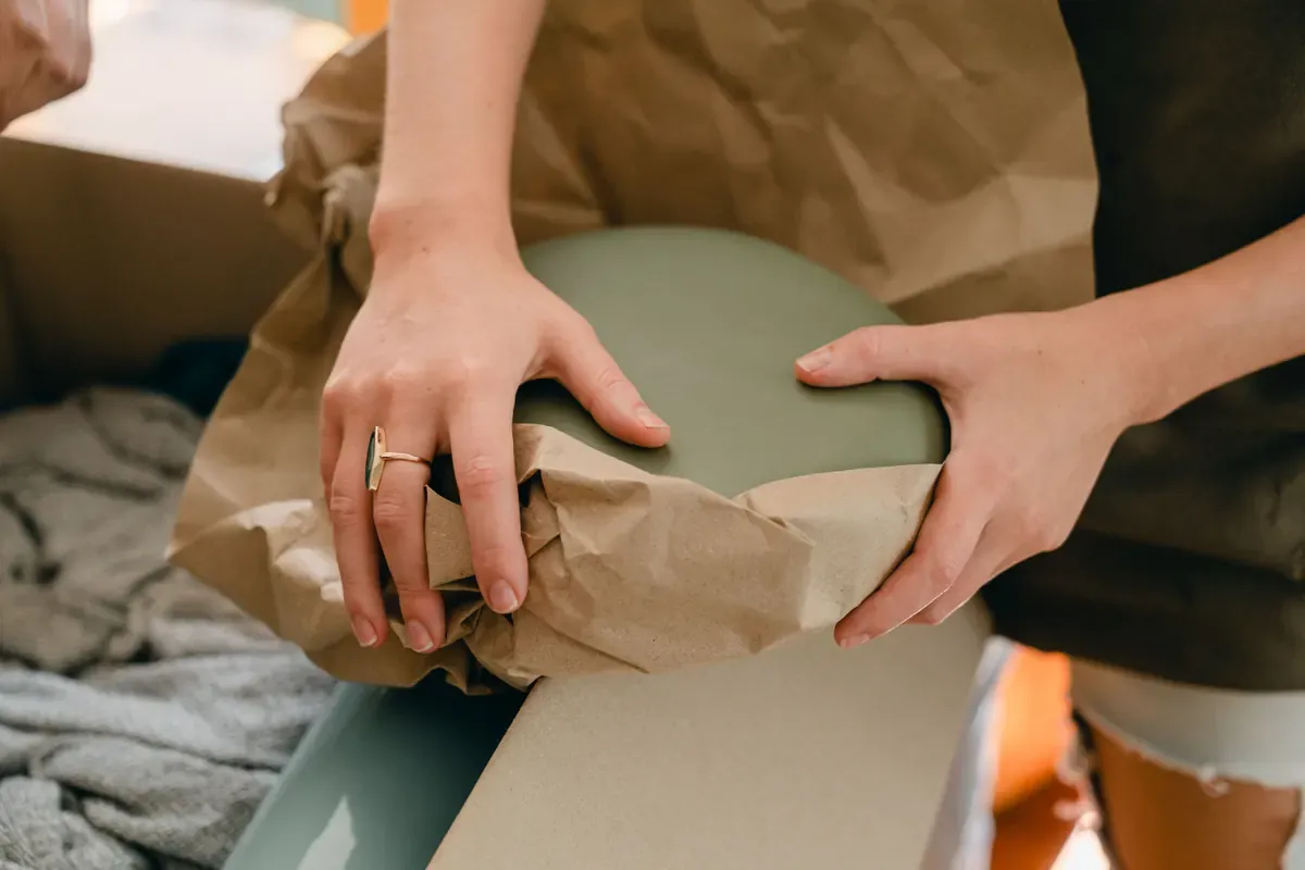 A person wraps a fragile ceramic item in packing paper before placing it in a box.