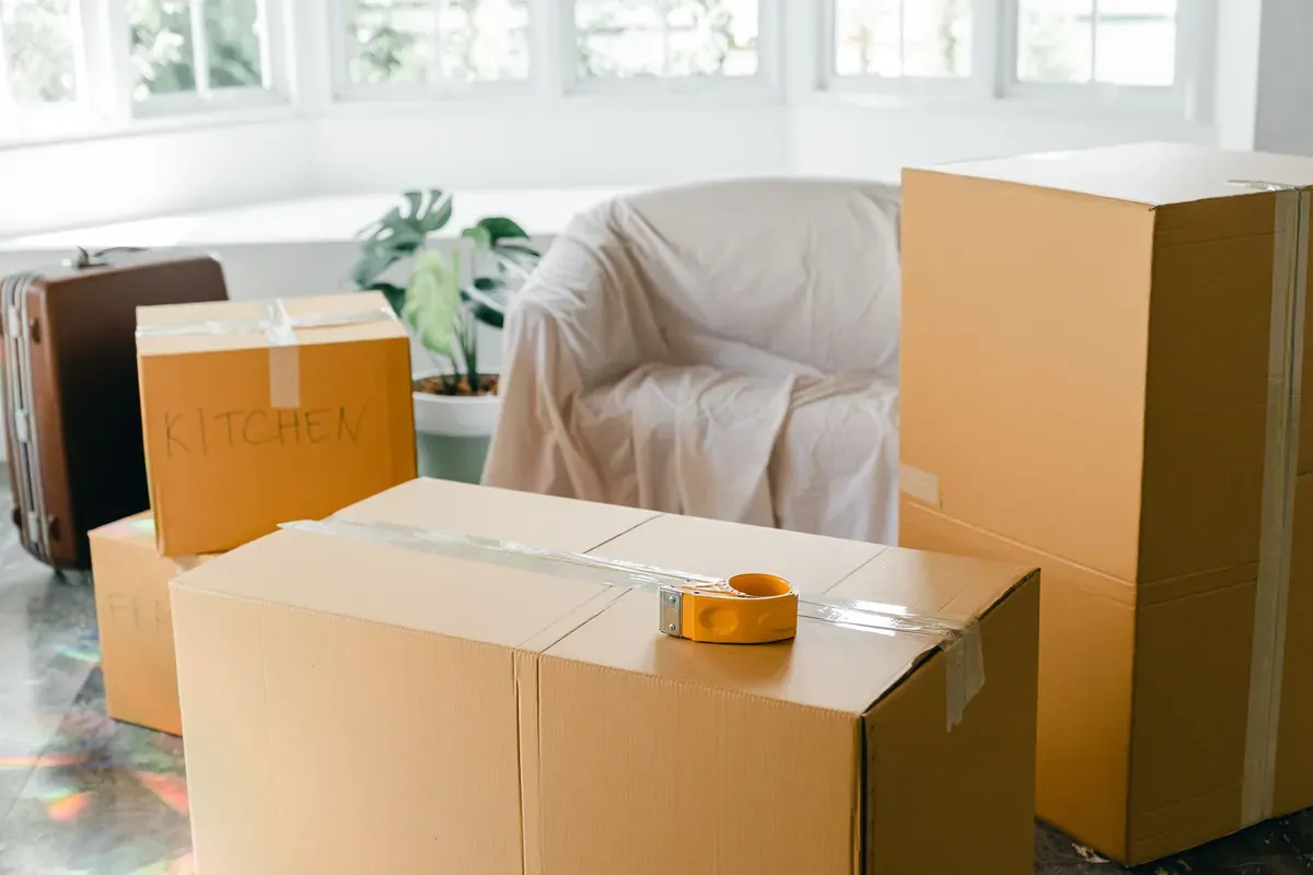 Packed moving boxes, including one labeled Kitchen, stand beside a covered sofa.
